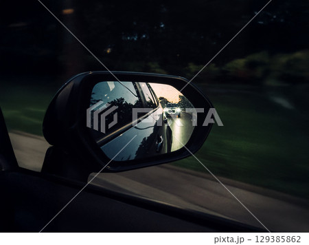 Close-up of a side mirror showing a car approaching from behind with headlights on during dusk or early evening. Motion blur, nighttime driving, driver safety, headlight glare risk Close-up of a side mirror showing a car approaching from behind with headlights on during dusk or early evening. Motion blur, nighttime driving, driver safety, headlight glare risk 129385862