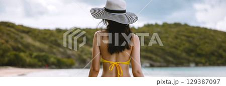 woman wearing  yellow bikini and  black and white hat is standing on  beach 129387097