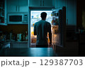 Man stands in front of open refrigerator, bright light illuminating his figure in a dark kitchen. Warm cozy environment in home, perfect for late night snacking. 129387703