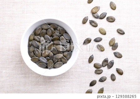 Styrian oil pumpkin seeds, in a white bowl, on linen. Dried, dark green and young fruits of the Styrian pumpkin, Cucurbita pepo var. styriaca, a variety of the common pumpkin, cultivated for seed oil. Styrian oil pumpkin seeds, in a white bowl, on linen. Dried, dark green and young fruits of the Styrian pumpkin, Cucurbita pepo var. styriaca, a variety of the common pumpkin, cultivated for seed oil. 129388265
