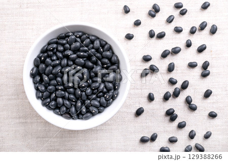 Black beans, in a white bowl, on linen. Dried variety of common beans, Phaseolus vulgaris, also known as black turtle beans, and caviar criollo. Type of bean with satiny black skin and white center. 129388266