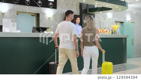 Man and woman going to hotel reception, where woman manager welcoming guests. Receptionists assisting visitors with checking-in. Travel and reception desk concept. Man and woman going to hotel reception, where woman manager welcoming guests. Receptionists assisting visitors with checking-in. Travel and reception desk concept. 129394384