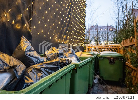 Green garbage bins with trash bags near Christmas tree and festive lights Green garbage bins with trash bags near Christmas tree and festive lights 129397121