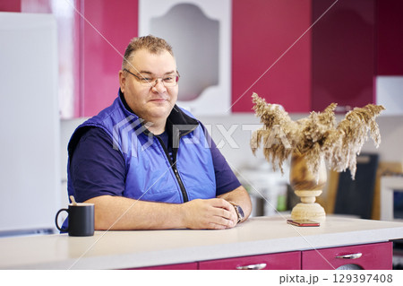 Body-positive European man with satisfied face sits in front of kitchen island. 129397408