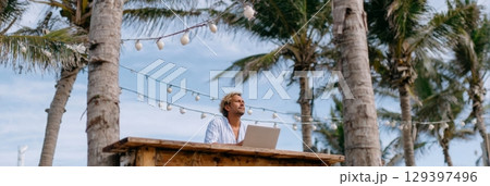Solo traveler working on laptop at wooden desk under palm trees, enjoying freedom and inspiration in a serene outdoor setting 129397496