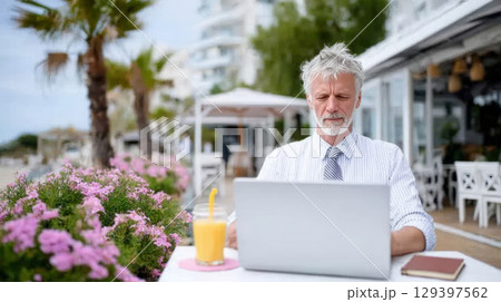 Senior man working remotely on laptop at outdoor cafe surrounded by vibrant flowers, enjoying the freedom of a flexible work environment 129397562