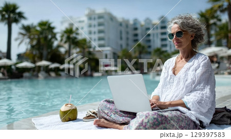 Senior woman working on laptop by the poolside, enjoying a tropical workcation with palm trees and a refreshing coconut drink nearby 129397583