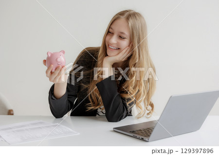 Young woman smiles while holding a piggy bank at her desk with a laptop and financial documents in a bright workspace Young woman smiles while holding a piggy bank at her desk with a laptop and financial documents in a bright workspace 129397896