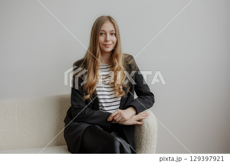 Woman in a black blazer and striped shirt poses confidently on a light-colored couch in a minimalistic indoor space 129397921