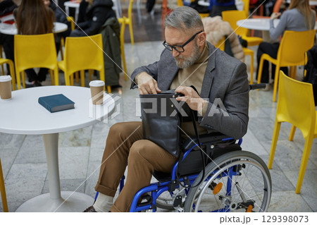 A stylish elderly man in a wheelchair thoughtfully organizes his bag in a busy cafe alone 129398073