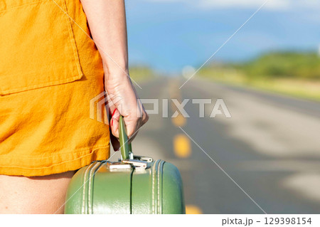 Woman's hand is holding a retro green suitcase and walking on a highway. 129398154