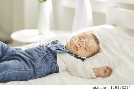 Adorable infant baby in denim overalls peacefully sleeping on white bed during calm daytime nap. Adorable infant baby in denim overalls peacefully sleeping on white bed during calm daytime nap. 129398214