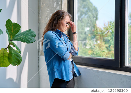 Mature tired, stressed woman standing at home near window Mature tired, stressed woman standing at home near window 129398300