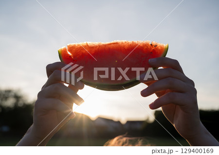 Juicy red watermelon slice in hand, on sunset park background 129400169