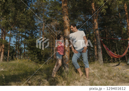father and daughter stand in a forest clearing near a tree next to a hammock and look at each other in summer, lifestyle 129401612