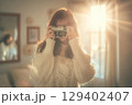 Woman Holding Camera in Cozy Setting with Warm Sunlight and Soft Background Elements 129402407