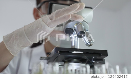 Female scientist wearing white protective gear analyzing test samples under microscope in contemporary research environment, close up of white gloved hand. Medicine and science concept 129402415