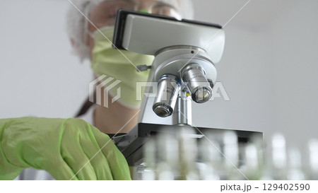 Female scientist wearing green mask and protective glasses using microscope in laboratory, doing scientific research. Medicine, healthcare and science concept 129402590