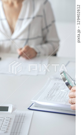 Professional accountants are working together, using a magnifying glass and a calculator, to audit financial reports in office, close up vertical view. Audit and taxes concept Professional accountants are working together, using a magnifying glass and a calculator, to audit financial reports in office, close up vertical view. Audit and taxes concept 129402612