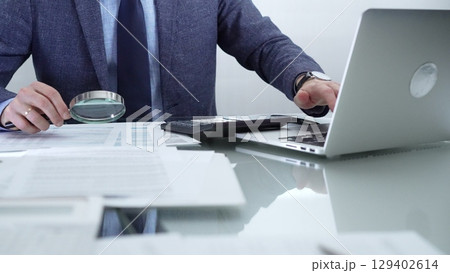 Businessman wearing blue suit is analyzing financial reports with magnifying glass and using calculator in a professional office setting. Low lighting, close-up view. Business people and audit concept Businessman wearing blue suit is analyzing financial reports with magnifying glass and using calculator in a professional office setting. Low lighting, close-up view. Business people and audit concept 129402614