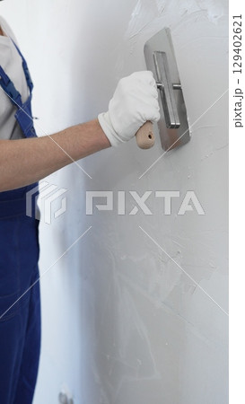 Professional construction worker wearing blue work overall is using a finishing trowel to carefully apply plaster on a wall, demonstrating expertise in home renovation and interior finishing Professional construction worker wearing blue work overall is using a finishing trowel to carefully apply plaster on a wall, demonstrating expertise in home renovation and interior finishing 129402621