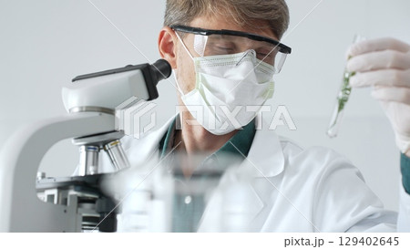 Adult male scientist in mask and gloves holding test tube with green plant sample, conducting laboratory research with a microscope. Medicine, health care and science concept 129402645