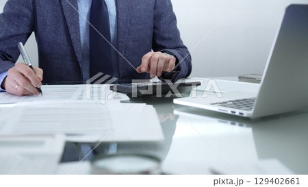 Businessman or accountant wearing blue suit is working with laptop, calculator and documents in office. Low lighting, close-up view. Business people, audit concept 129402661