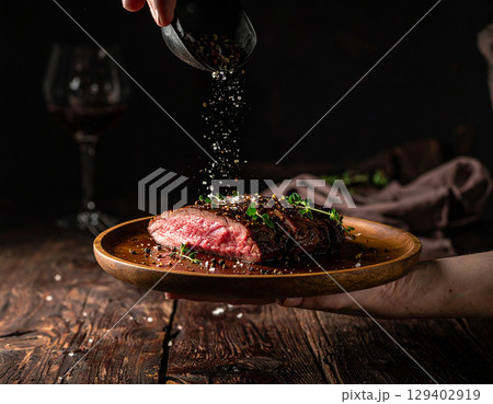 AI generated close-up of a perfectly cooked medium rare steak resting on a hand-held plate, garnished with falling black pepper and salt in an intimate dimly-lit dining setting 129402919