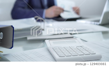 Calculator, pen, laptop and documents are lying on accountant's desk. Businessman wearing blue suit is working with documents on the background. Business people, audit concept Calculator, pen, laptop and documents are lying on accountant's desk. Businessman wearing blue suit is working with documents on the background. Business people, audit concept 129403081