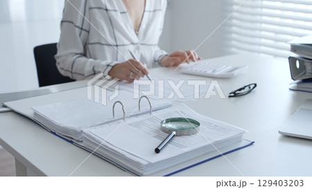 Female accountant is working with financial documents, using calculator and taking notes, with a magnifying glass and a folder on a white table in a bright office. Audit and taxes in business 129403203