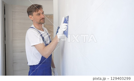 Man construction worker wearing protective white gloves and blue construction coveralls, is sanding white wall with blue hand block, preparing smooth surface during interior home renovation project Man construction worker wearing protective white gloves and blue construction coveralls, is sanding white wall with blue hand block, preparing smooth surface during interior home renovation project 129403299
