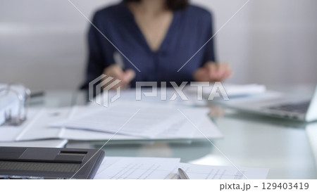 Black calculator, pen and documents are lying on accountant's desk. Businesswoman wearing blue dress is working with documents on the background. Business and audit concept Black calculator, pen and documents are lying on accountant's desk. Businesswoman wearing blue dress is working with documents on the background. Business and audit concept 129403419