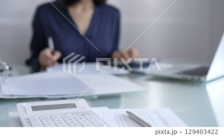 White calculator, pen and documents are lying on accountant's desk. Businesswoman wearing blue dress is working with documents on the background. Business and audit concept White calculator, pen and documents are lying on accountant's desk. Businesswoman wearing blue dress is working with documents on the background. Business and audit concept 129403422