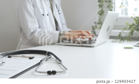 Stethoscope over a medical records are lying on the desk opposite the doctor is typing on a laptop with a in a medical office, suggesting online healthcare or telemedicine services. Medicine concept 129403427