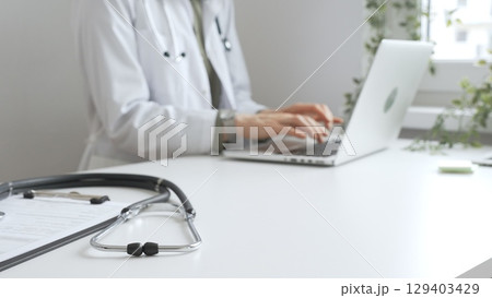 Stethoscope over a medical records are lying on the desk opposite the doctor is typing on a laptop with a in a medical office, suggesting online healthcare or telemedicine services. Medicine concept Stethoscope over a medical records are lying on the desk opposite the doctor is typing on a laptop with a in a medical office, suggesting online healthcare or telemedicine services. Medicine concept 129403429