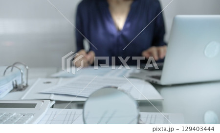 Magnifying glass, white calculator, pen and documents are lying on accountant's desk. Businesswoman wearing blue dress is working with documents on the background. Business and audit concept 129403444