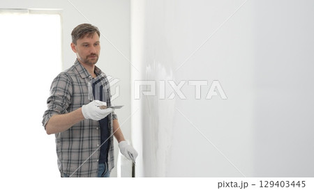 Construction worker wearing gloves and using a putty knife to plaster a white wall, performing home renovation and improvement tasks with precision Construction worker wearing gloves and using a putty knife to plaster a white wall, performing home renovation and improvement tasks with precision 129403445