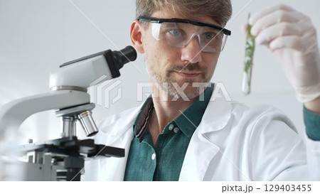 Adult male scientist in mask and gloves holding test tube with green plant sample, conducting laboratory research with a microscope. Medicine, healthcare and science concept 129403455