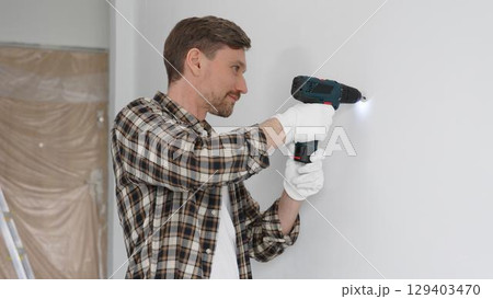 Man construction worker in checked shirt and safety gloves is screwing a screw into the wall using a cordless screwdriver, carrying out a careful home renovation. Horizontal portrait view 129403470