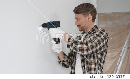 Male construction worker in checked shirt and safety gloves is screwing a screw into the wall using a cordless screwdriver, carrying out a careful home renovation. Portrait view 129403574