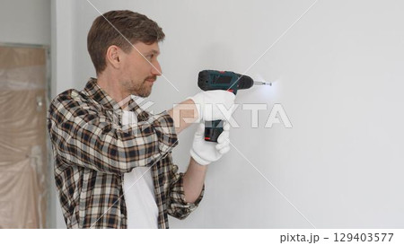 Middle aged man wearing checked shirt and white gloves, is drilling hole in a wall with cordless drill during home renovation work. Portrait view 129403577