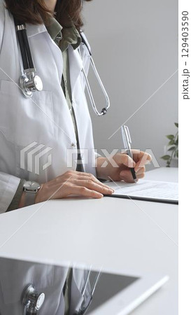 Professional female doctor wearing white lab coat and stethoscope documenting patient information on clipboard near laptop in clinic workspace. Medicine and health care concept 129403590