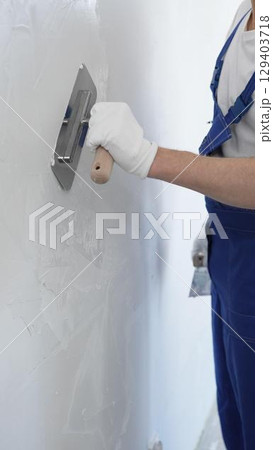 Professional construction worker wearing blue work overall is using a finishing trowel to carefully apply plaster on a wall, demonstrating expertise in home renovation 129403718