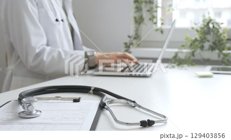 Stethoscope and medical records are on the desk opposite the doctor is typing on a laptop with a in a medical office, suggesting online healthcare or telemedicine services. Medicine concept 129403856