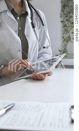 Doctor woman in lab coat and stethoscope using digital tablet at desk in medical office, accessing online patient information. Medicine and health care concept 129403860
