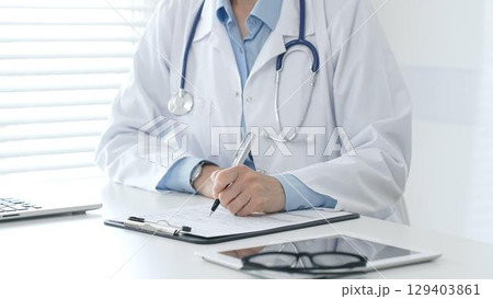 Female doctor documents patient information while working with digital devices at minimalist white workstation in medical office. Medicine and healthcare concept 129403861
