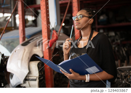 Confident african american female worker holding blue folder while inspecting car parts warehouse. Concept of automotive maintenance, quality control industrial service. 129403867