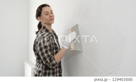Woman construction professional wearing checkered shirt is smoothing white wall surface, applying putty with professional spatula technique, showcasing skilled interior renovation work 129403880
