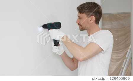 Middle aged male worker wearing white t-shirt and protective gloves, is drilling hole in a wall with cordless drill during home renovation work. Portrait view 129403909