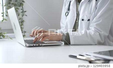 Doctor woman wearing a stethoscope and lab coat is diligently typing on a laptop, showcasing efficient healthcare management in a modern medical office. Medicine and health care 129403958